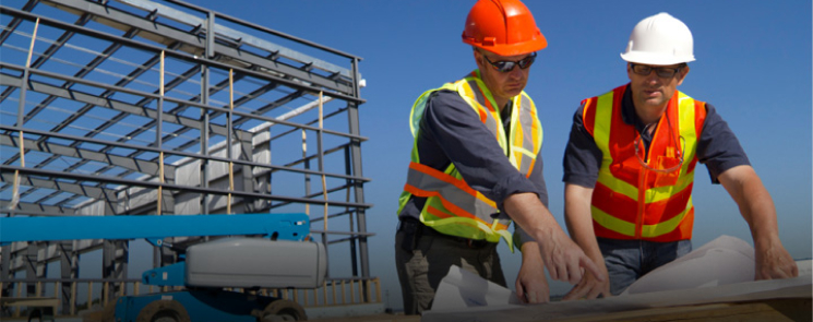 Two men wearing helmets and safety gear reviewing blueprints at a construction site with a building under construction in the background.